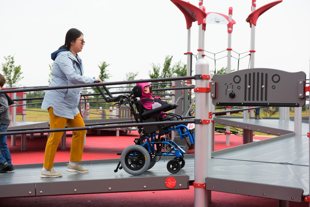 young girl in wheelchair being pushed up playground ramp by caregiver