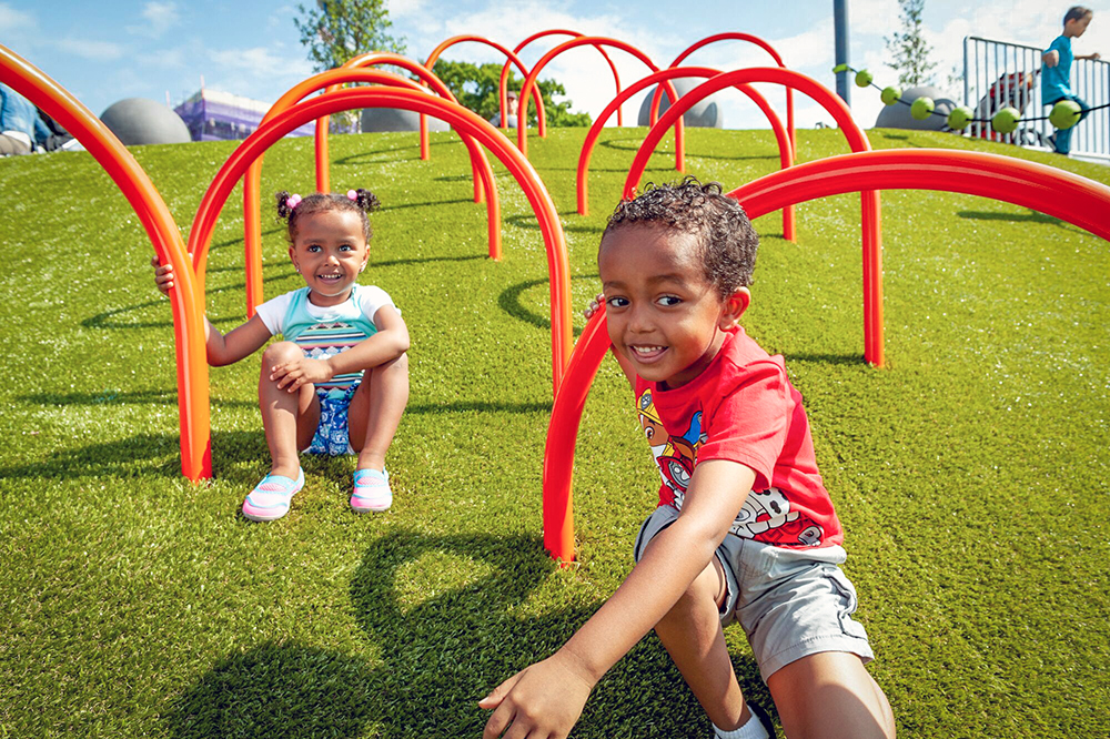 Two children sliding down turf coloured hillside playgrounds through orange loops