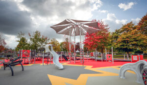a grey accessible playground with slides and shade on a colourful rubber surface