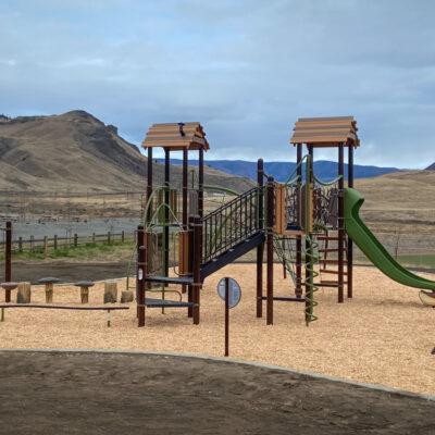 Green and brown playground with wood accents and swings on wood fibre surfacing with mountains in background