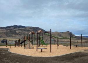 Green and brown playground with wood accents and swings on wood fibre surfacing with mountains in background