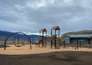 Green and brown playground with wood accents and swings on wood fibre surfacing with mountains in background