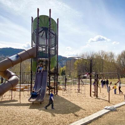Green and brown playground tower with horizontal and vertical logs and swings on wood fibre surfacing