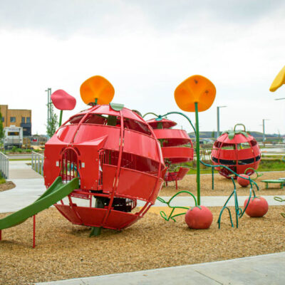 A playground featuring large red fruit-shaped climbing structures and tall yellow and red flower sculptures on a mulch surface, with pavements and buildings in the background.