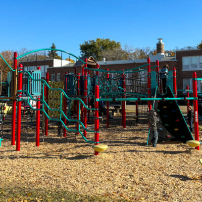 Outdoor playground with red and green climbing frames, slides, and ladders on wood chippings, with a school building and children playing in the background.